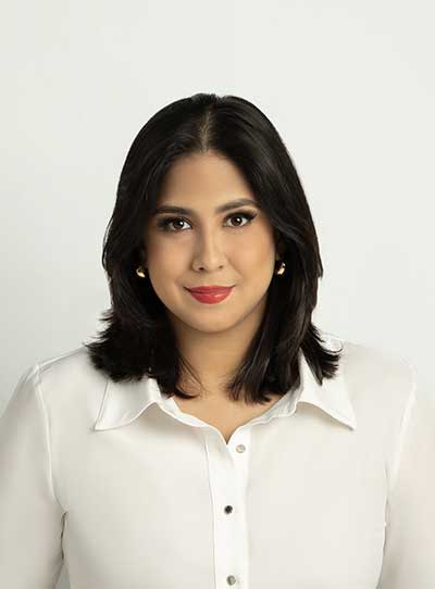 A Latina woman with black hair, red lipstick, makeup on, and a beige-colored button-down blouse; she has a fair light skin tone. She is looking straight ahead.