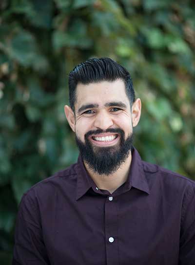 A young man of about 30 with a black beard, smiling, with thick eyebrows, pointed ears, and a casual violet shirt, against a background of green vegetation.