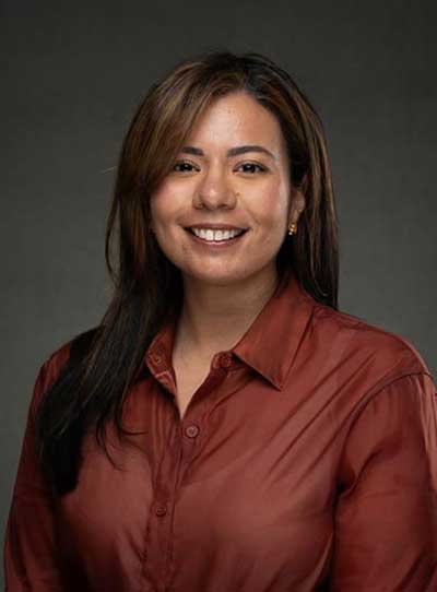 A dark-complexioned woman with long brown hair, smiling, wearing a burgundy blouse, against a dark grayish background.